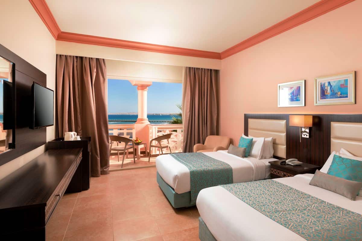 A hotel room with two beds, a TV, a desk, and a view of the sea.
