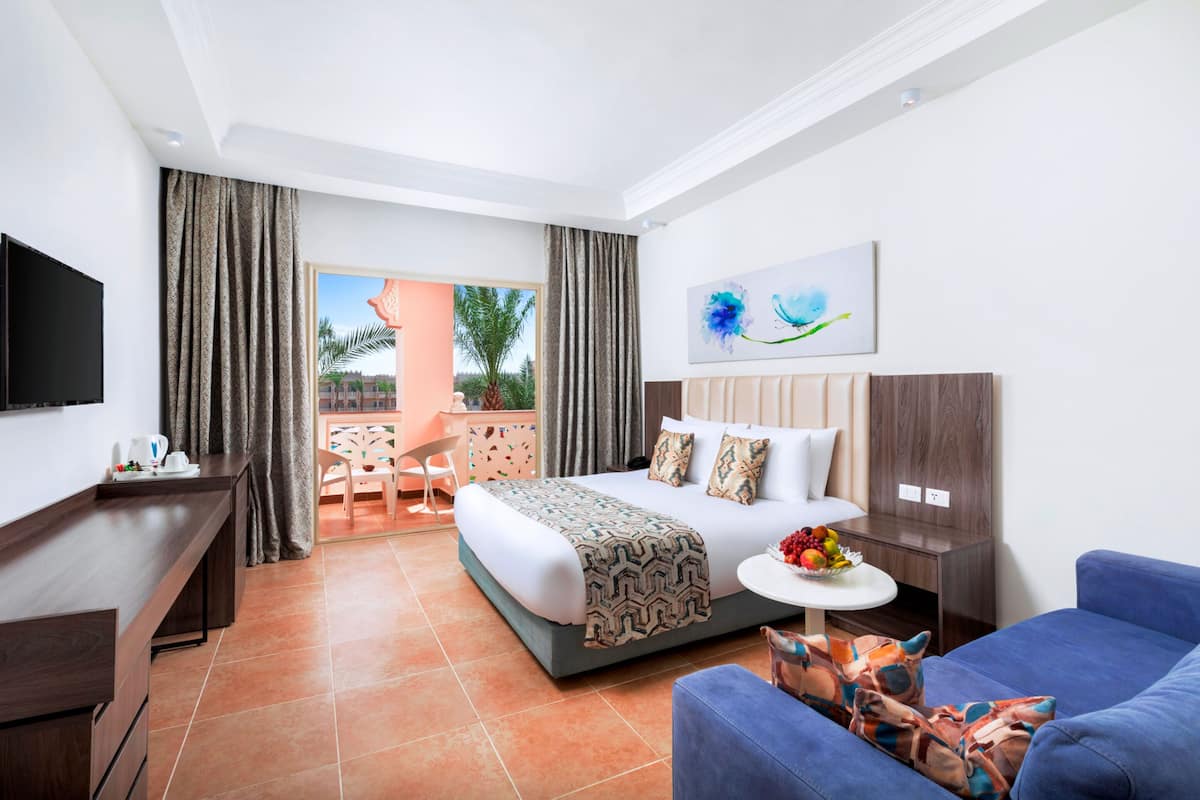 A hotel room with a bed, a TV, a desk, a chair, a small table with fruit, and a balcony with a view of palm trees.
