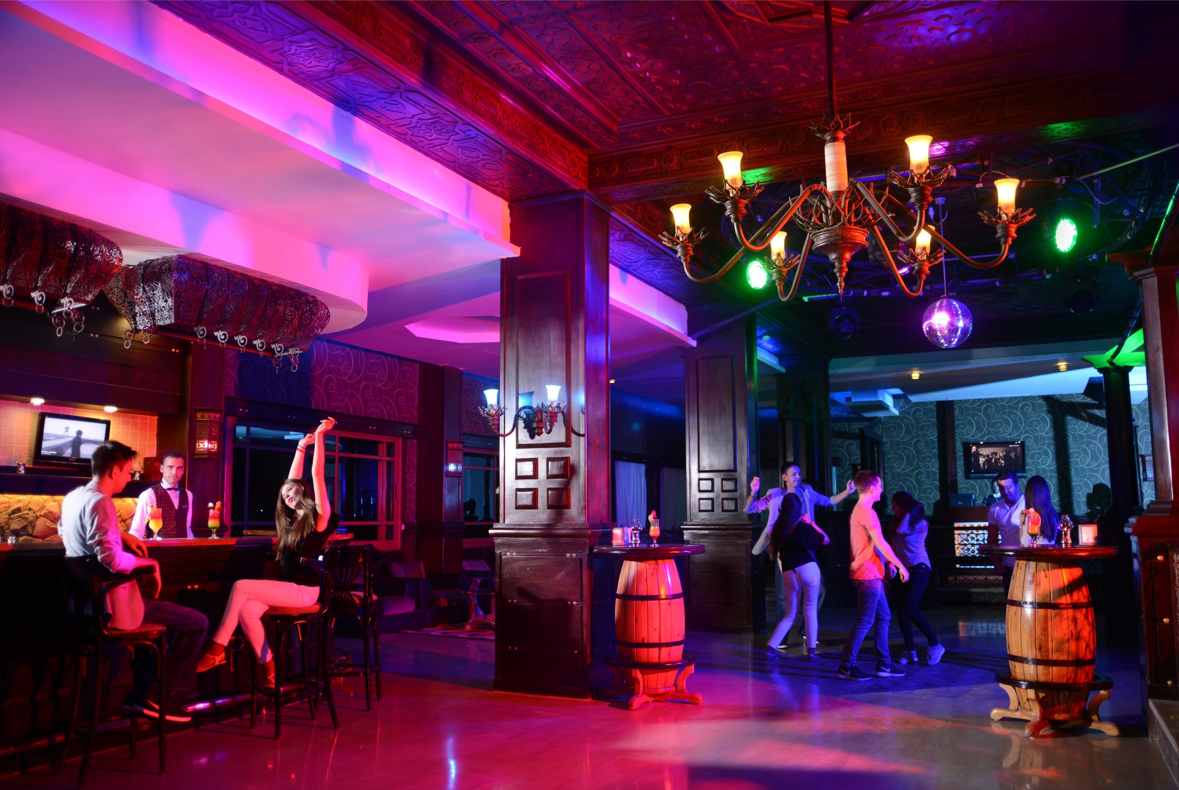 Een bar met houten toonbanken, krukken en een kroonluchter. Mensen maken er gezellig praatjes en dansen.