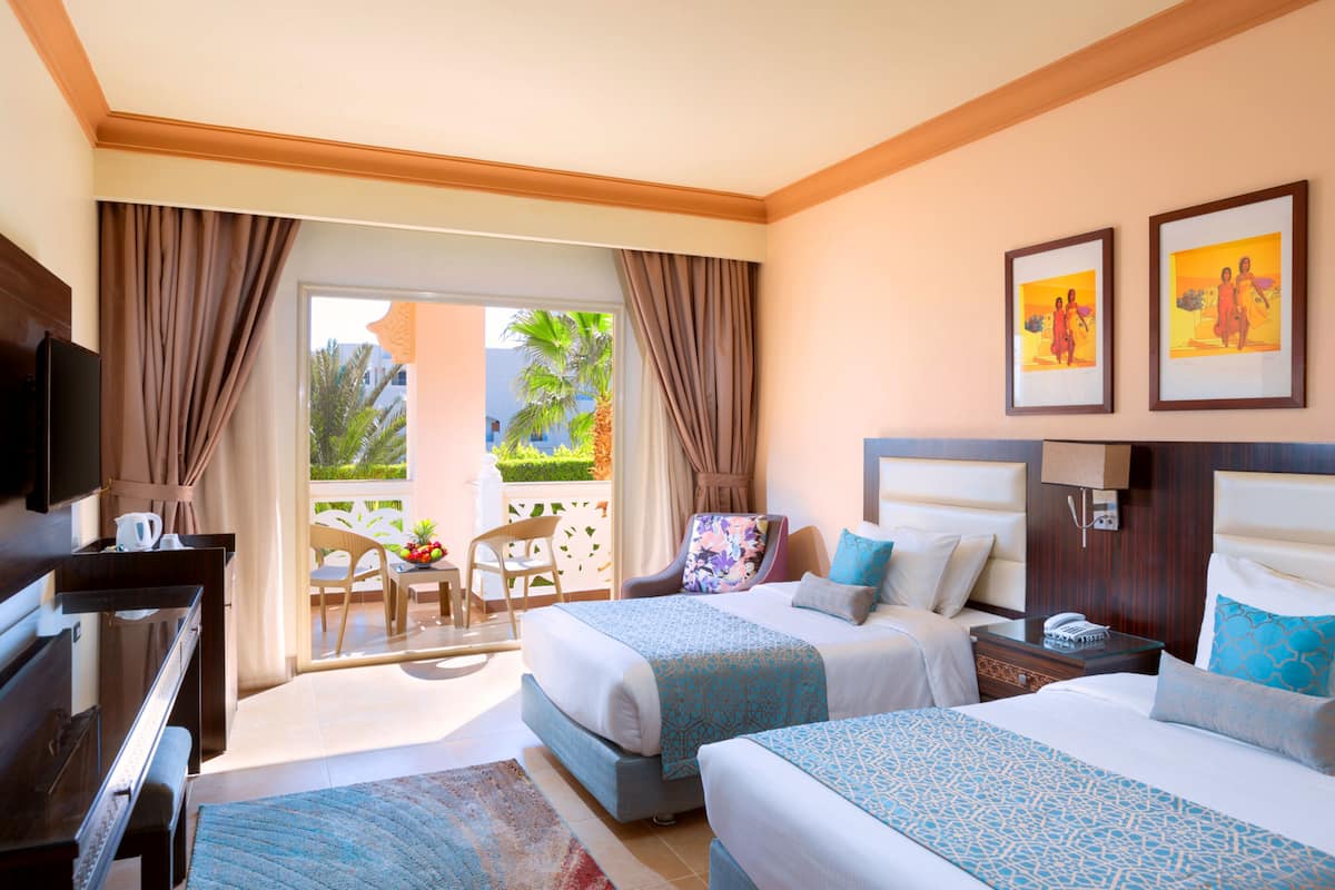 A hotel room with two beds, a desk, a TV, and a balcony with a chair and table.