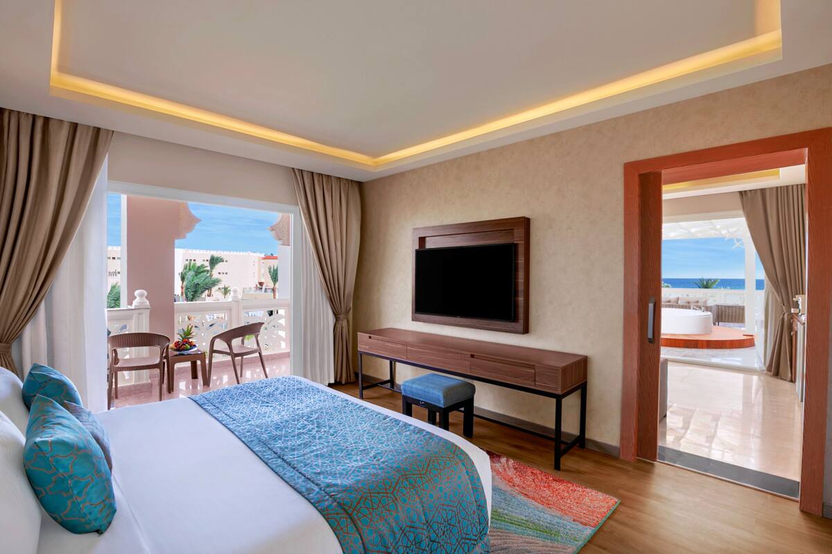A hotel room with a bed, a desk, a TV, and a balcony with a view of the sea.