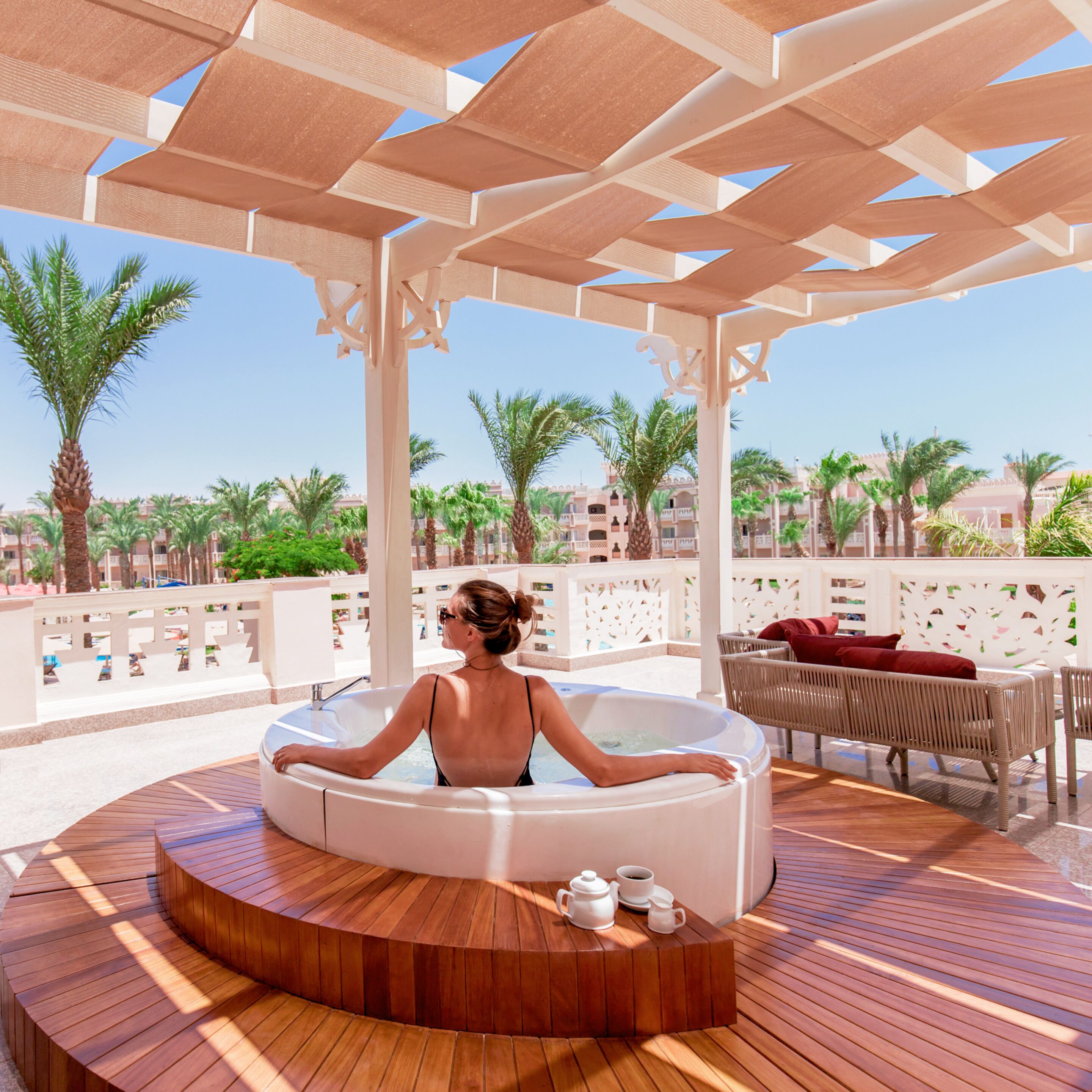A woman relaxing in a hot tub on a wooden deck with a view of palm trees and buildings.