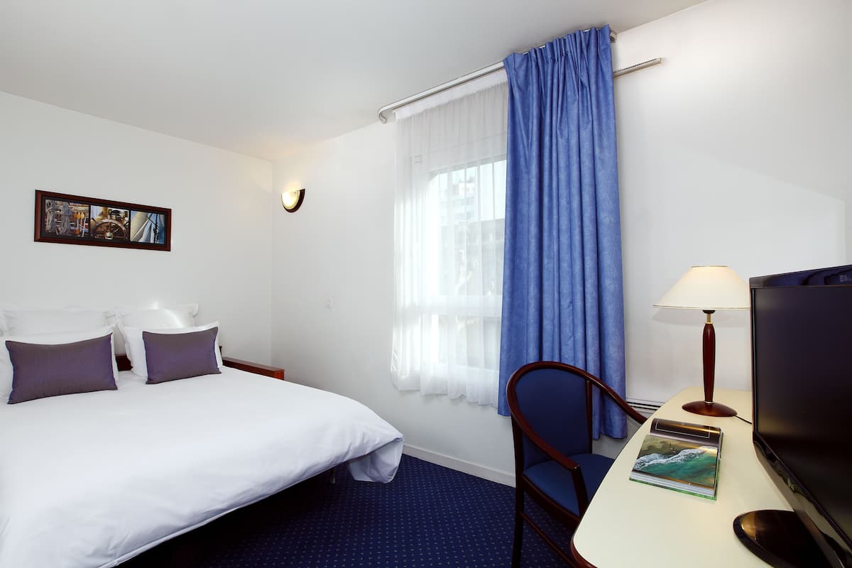 Una habitación de hotel con cama, escritorio con lámpara, televisión y ventana con cortinas.