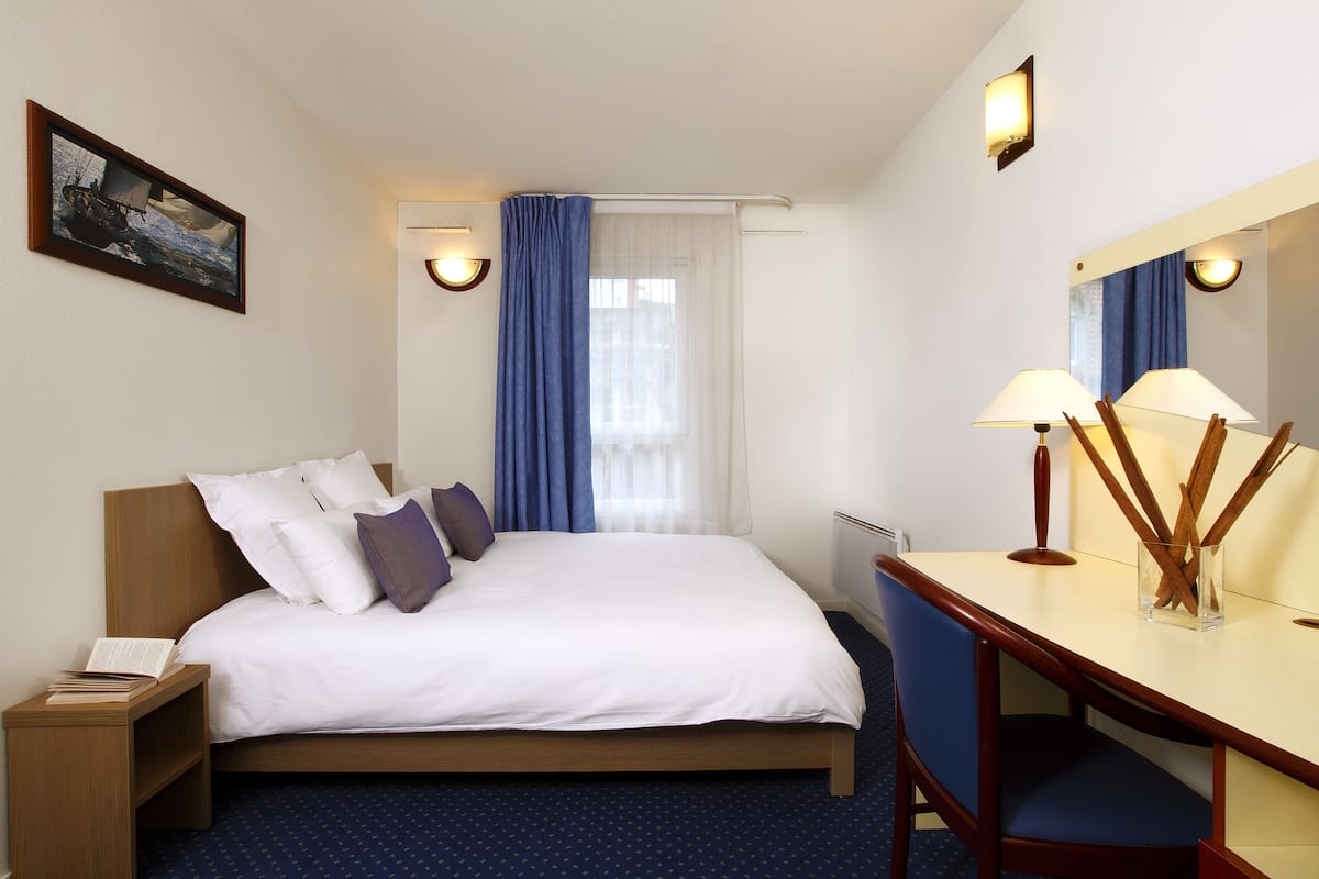 Una habitación de hotel con cama, escritorio con silla, mesita de noche, espejo y ventana con cortinas.