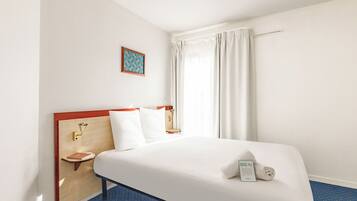 Habitación de hotel con cama, mesita de noche, lámpara de pared, ventana con cortinas y un cuadro en la pared.