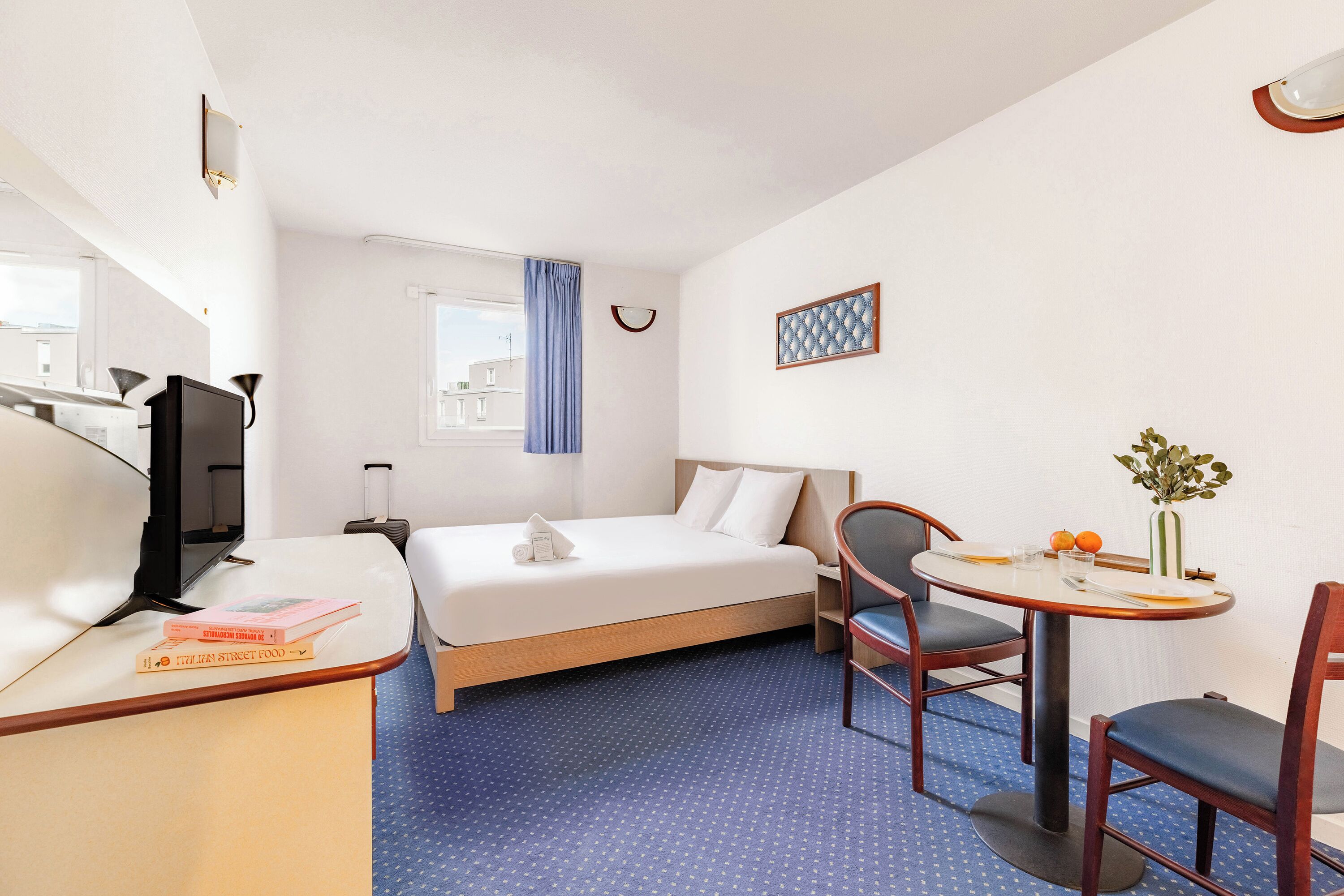 A hotel room with a bed, a desk with a chair, a small table, and a window with curtains.