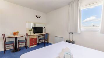A hotel room with a bed, a desk with a computer, a chair, a small table with fruit, a mirror, a window with curtains, and a radiator.