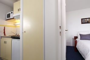 A compact hotel room with a kitchenette, a bed with white bedding, and a small bedside table.