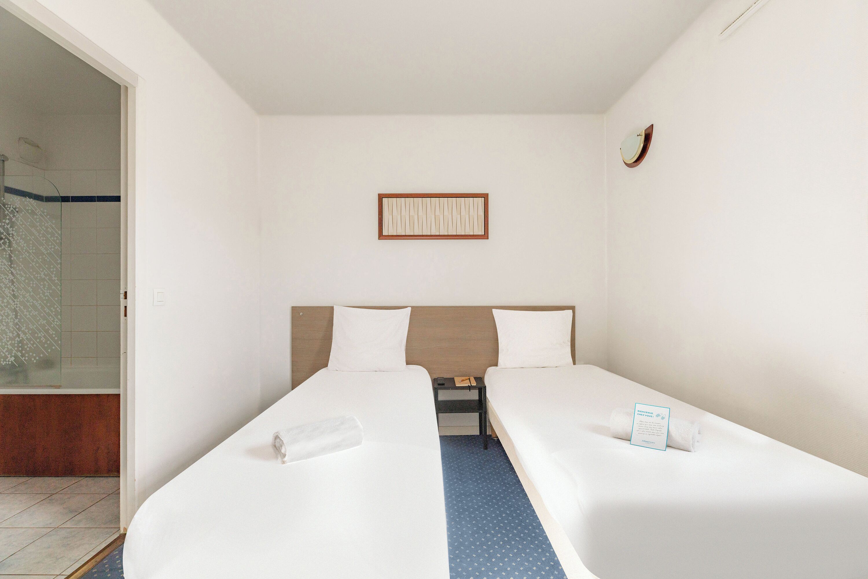 Habitación de hotel con dos camas individuales, una mesita de noche pequeña con lámpara, cabecero de madera, un cuadro enmarcado en la pared y zona de ducha con puerta de cristal.