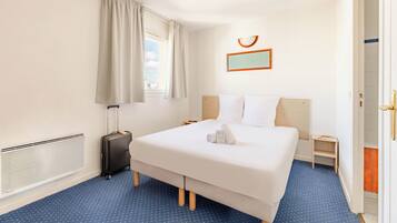 A hotel room with a bed, a suitcase, a radiator, a window with curtains, a nightstand, and a door.