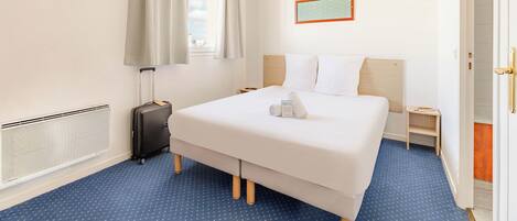 A hotel room with a bed, a suitcase, a radiator, a window with curtains, a nightstand, and a door.