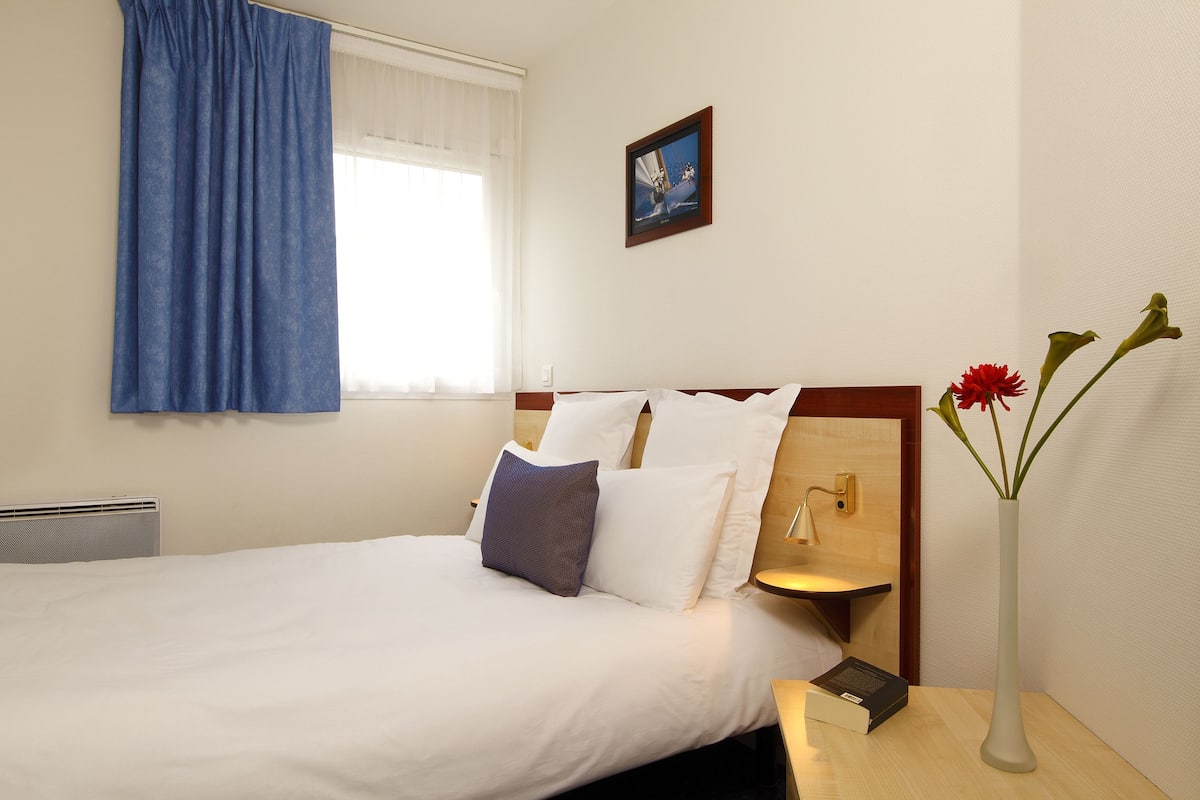 Una cama bien hecha con almohadas blancas y azules, una mesita de noche con un libro y un jarrón con flores, y una ventana con cortinas azules.