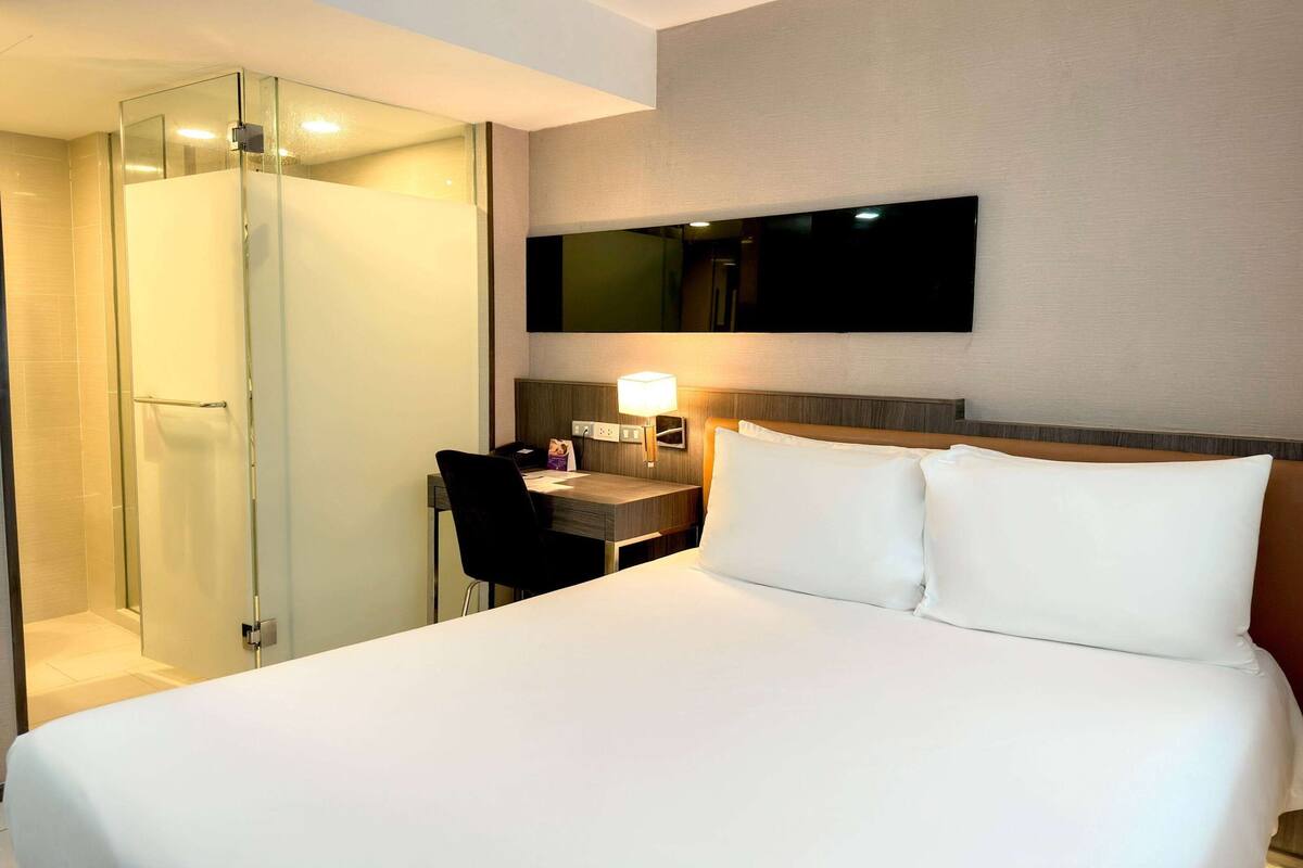 Habitación de hotel con cama, escritorio, silla, ducha y televisor montado en la pared.