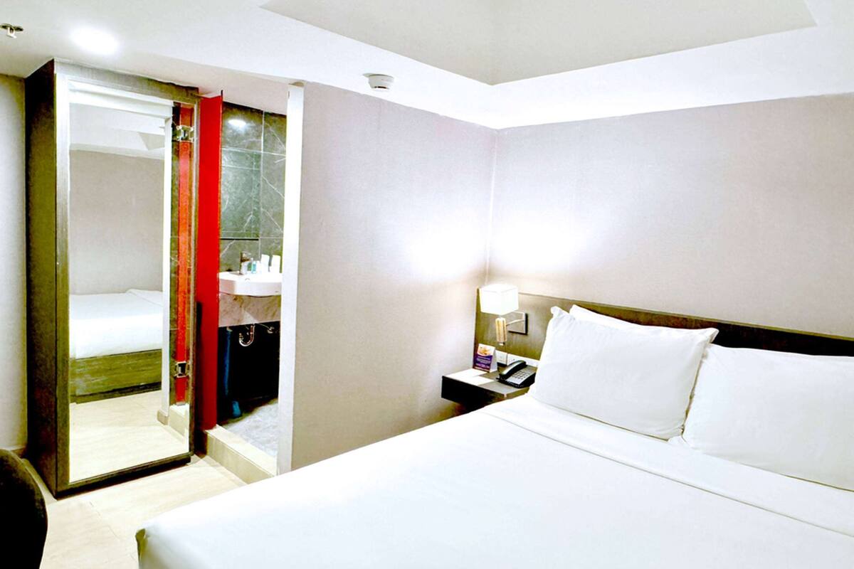 Habitación de hotel con cama, mesita de noche con lámpara, espejo y baño visible a través de una puerta abierta.