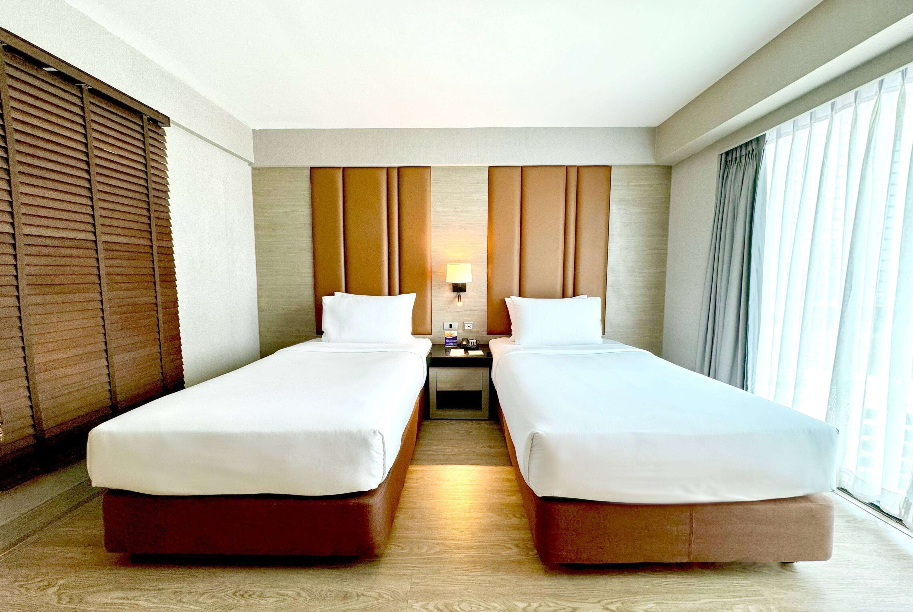 A hotel room with two single beds, a nightstand with a lamp, and a window with curtains.