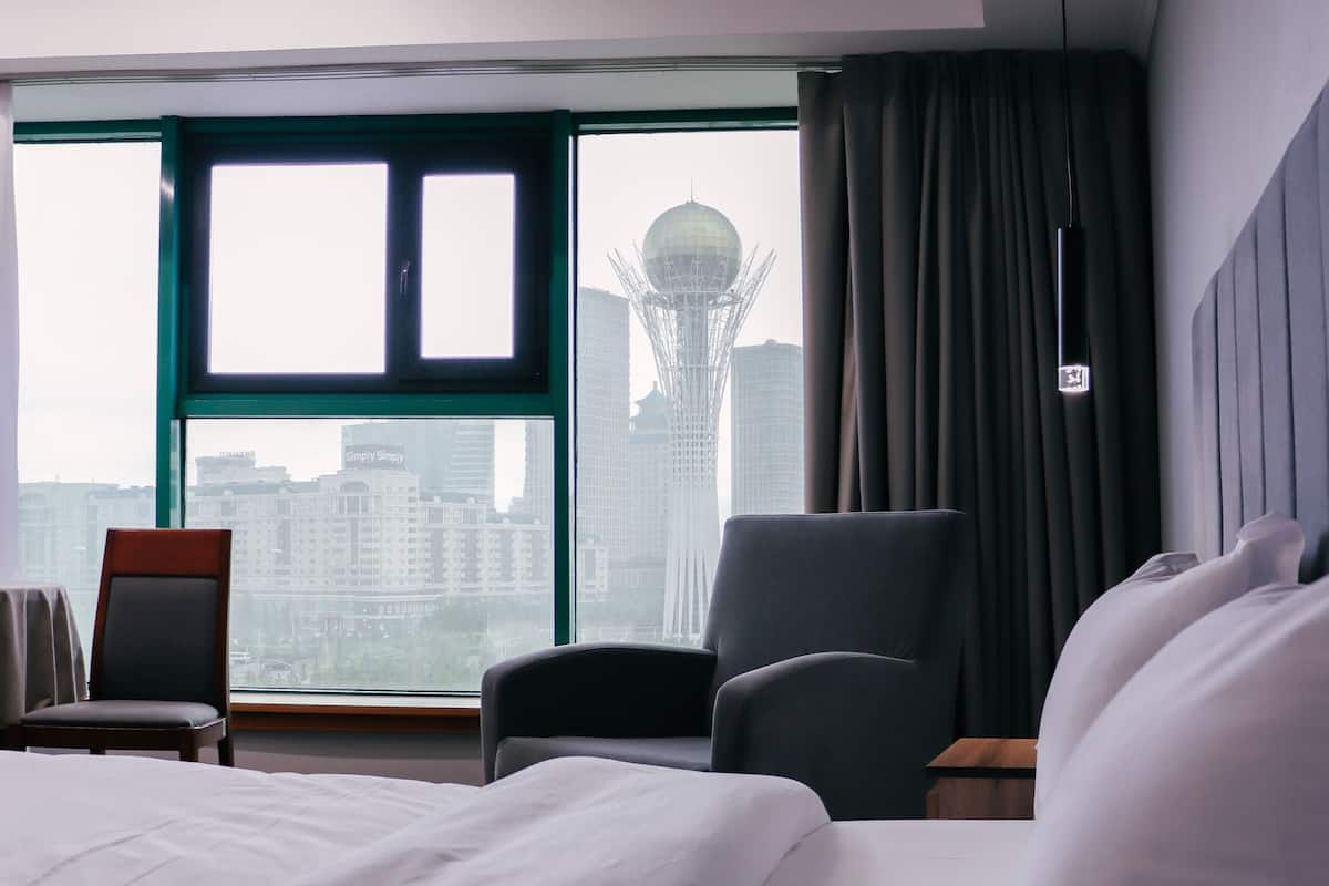 A hotel room with a large bed, a chair, a desk, and a view of a cityscape through a large window.