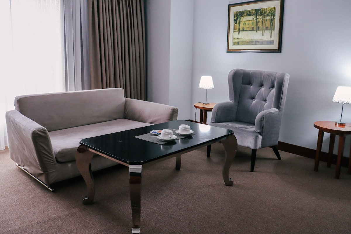 A modern hotel room with a grey sofa, a black coffee table with two white cups, a grey armchair, and two small wooden side tables with lamps.