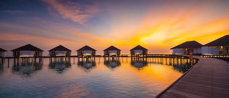 Overwater bungalows connected by a wooden walkway over calm water, with a sunset in the background.