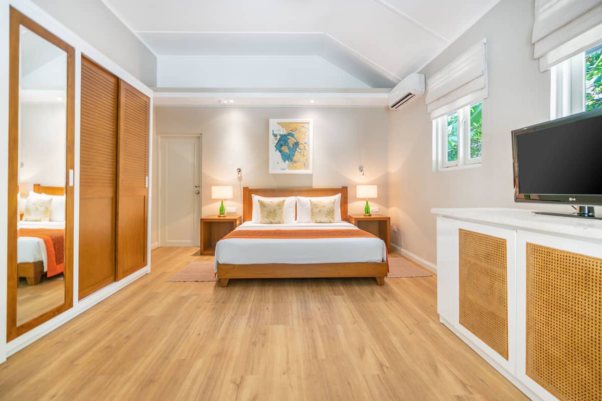 A modern bedroom with a large bed, wooden wardrobe, bedside tables with lamps, a flat-screen TV, and a large window with a view of greenery.