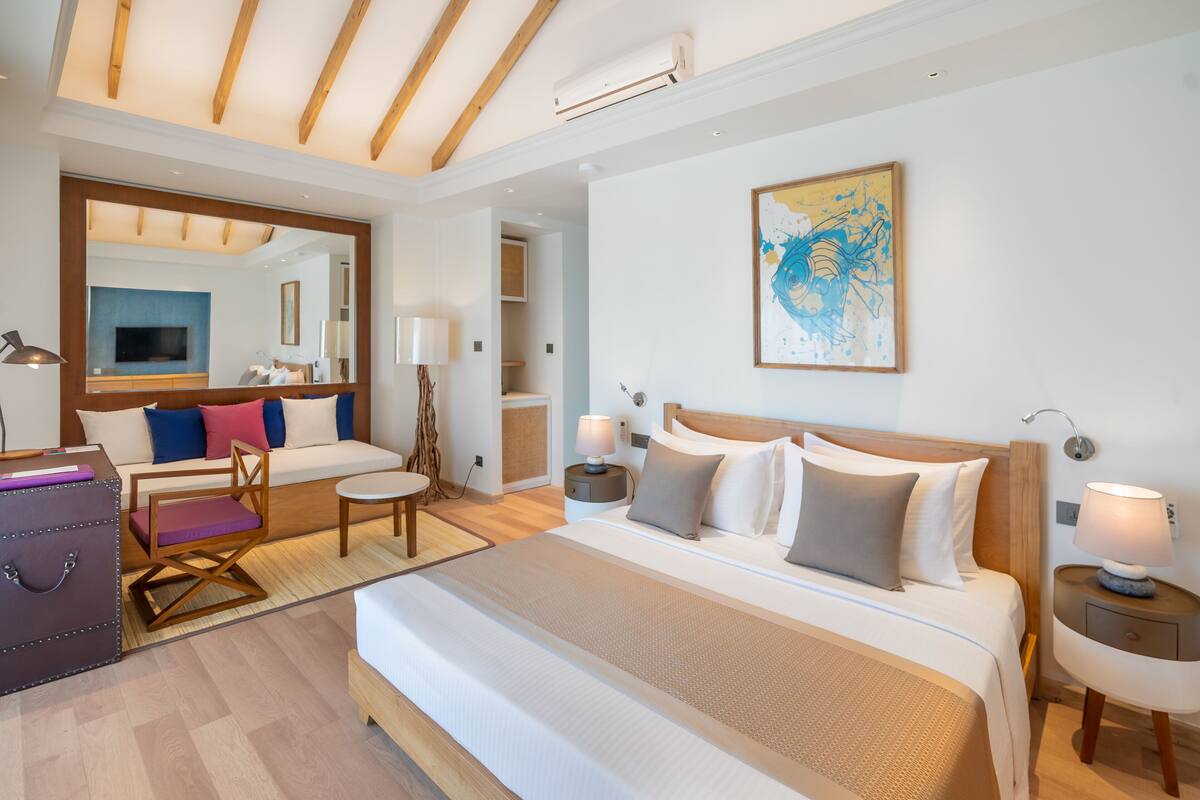 A modern hotel room with a large bed, a sitting area with a sofa and armchairs, a dining table, and a large mirror.