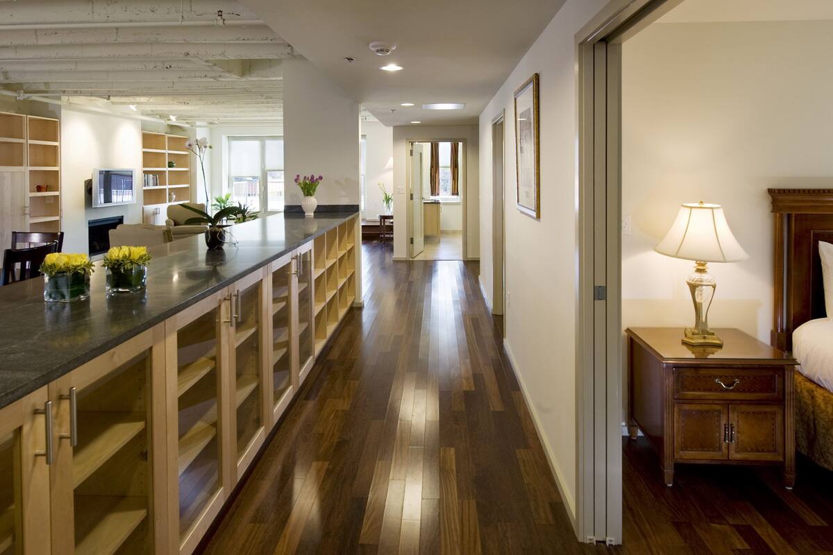 A modern hotel corridor with wooden flooring, a kitchen area with a counter and cabinets, and a bedroom with a nightstand and lamp.