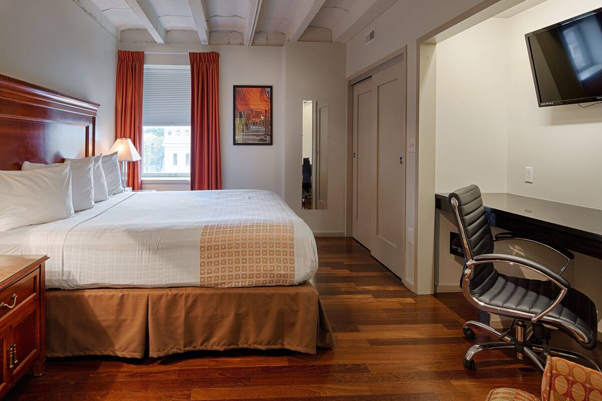A hotel room with a large bed, wooden headboard, a desk with a lamp, a chair, and a TV mounted on the wall.