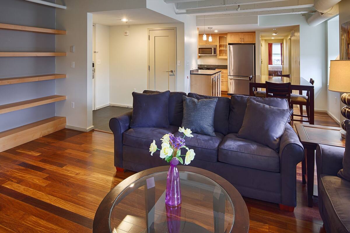 A modern living room with a dark sofa, a glass coffee table with flowers, wooden flooring, and a kitchen in the background.