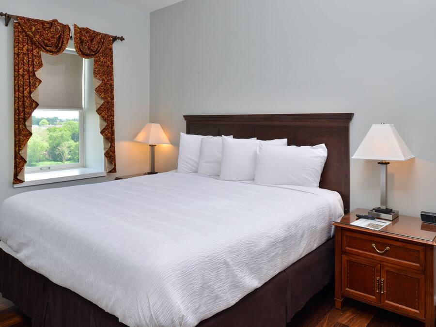 A neatly made bed with white linens, a wooden nightstand with a lamp, and a window with curtains.