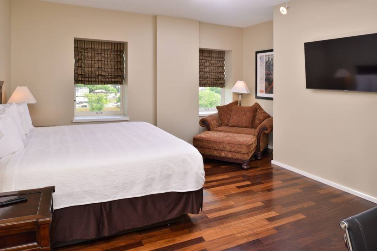A hotel room with a large bed, two windows with blinds, a chair, a flat-screen TV, and a wooden floor.
