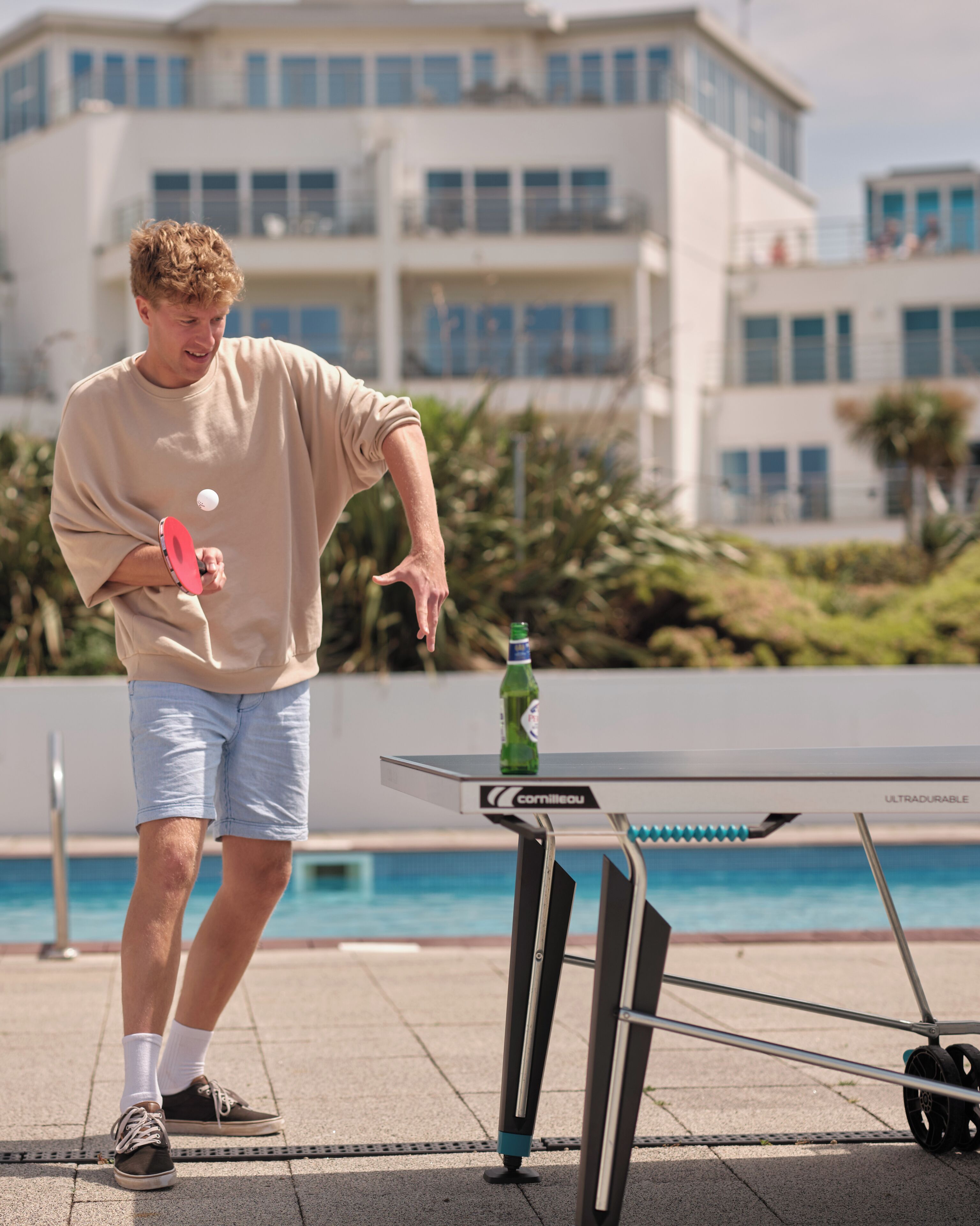 Una persona jugando ping pong al aire libre, con una mesa y una botella cerca.