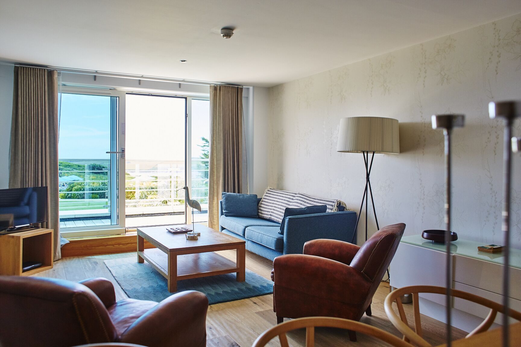 A modern hotel room with a balcony, a sofa, a coffee table, and leather chairs.