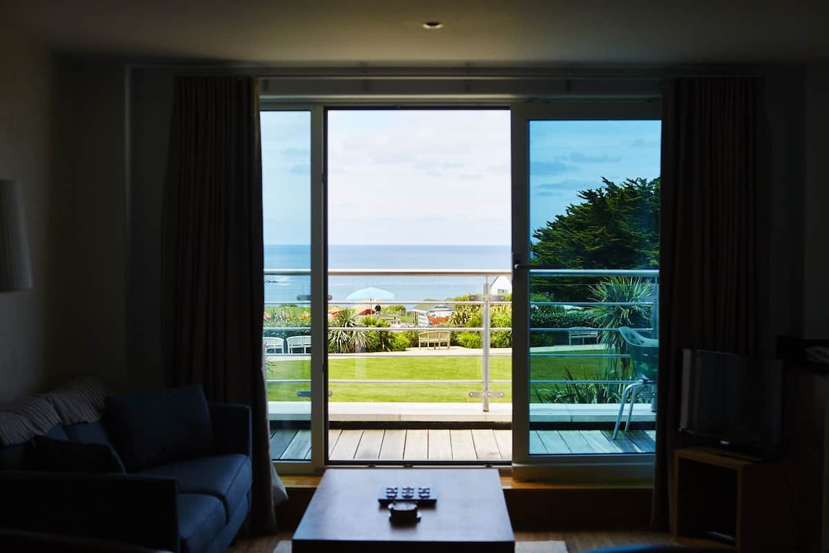 Habitación con sofá, una mesa de centro y televisor. Cuenta con un amplio ventanal corredizo que da a un balcón con vista al mar y un jardín.