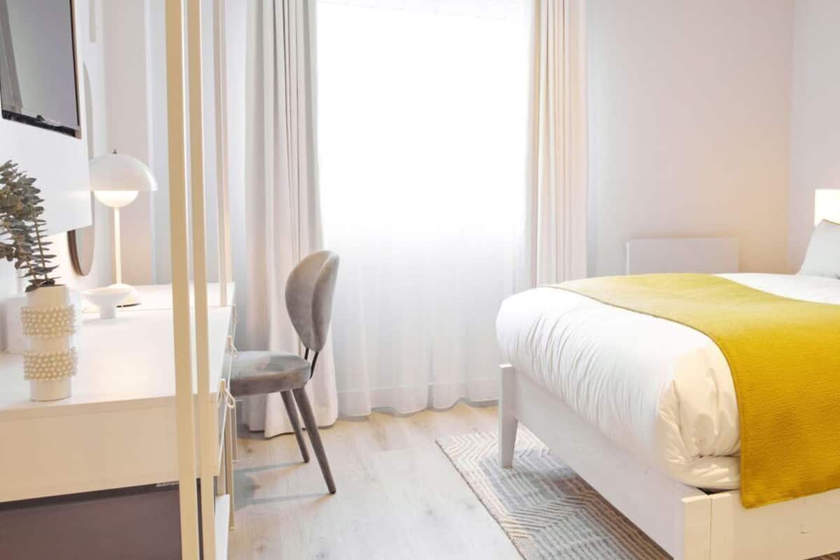 Una habitación de hotel con cama, mesita de noche, lámpara, silla y ventana con cortinas.
