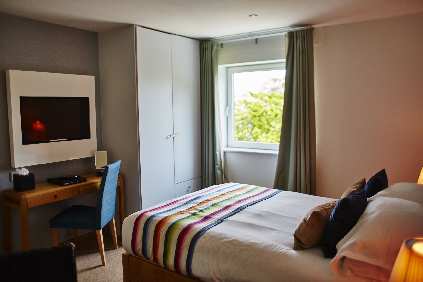 A hotel room with a bed, a desk with a computer, a TV, and a window with curtains.