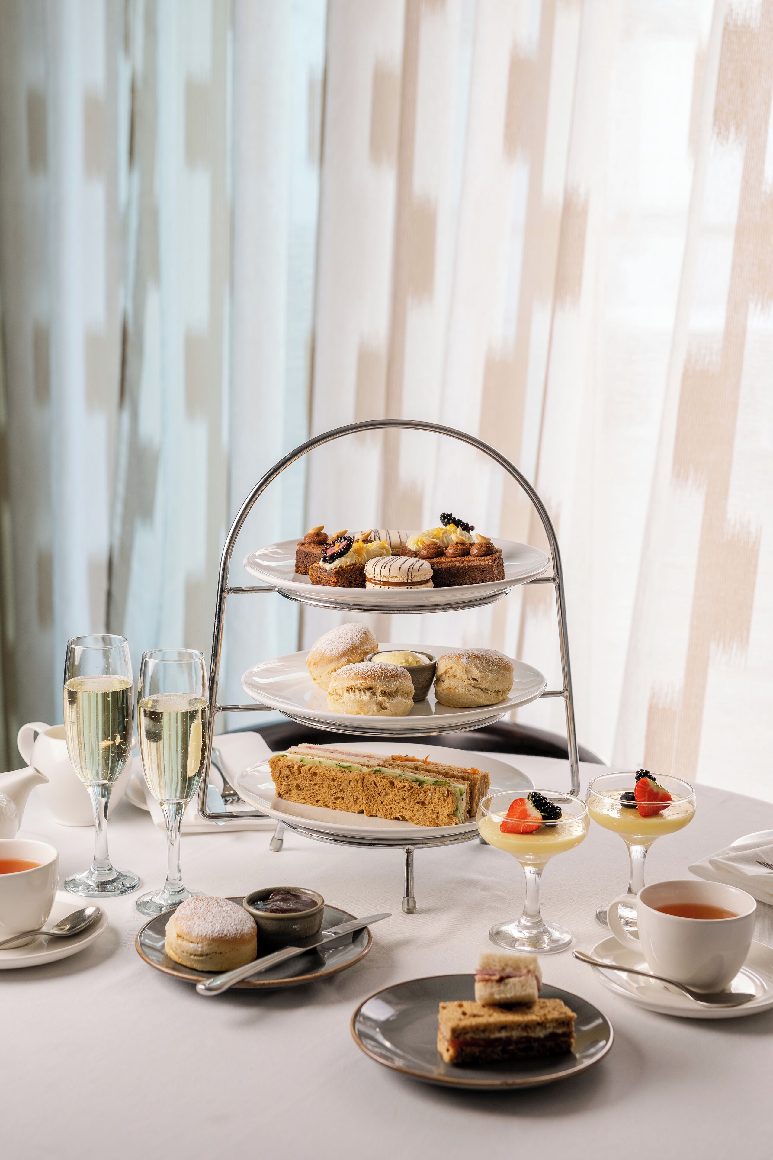 A three-tiered dessert stand with various pastries and sandwiches, accompanied by glasses of champagne and cups of tea.