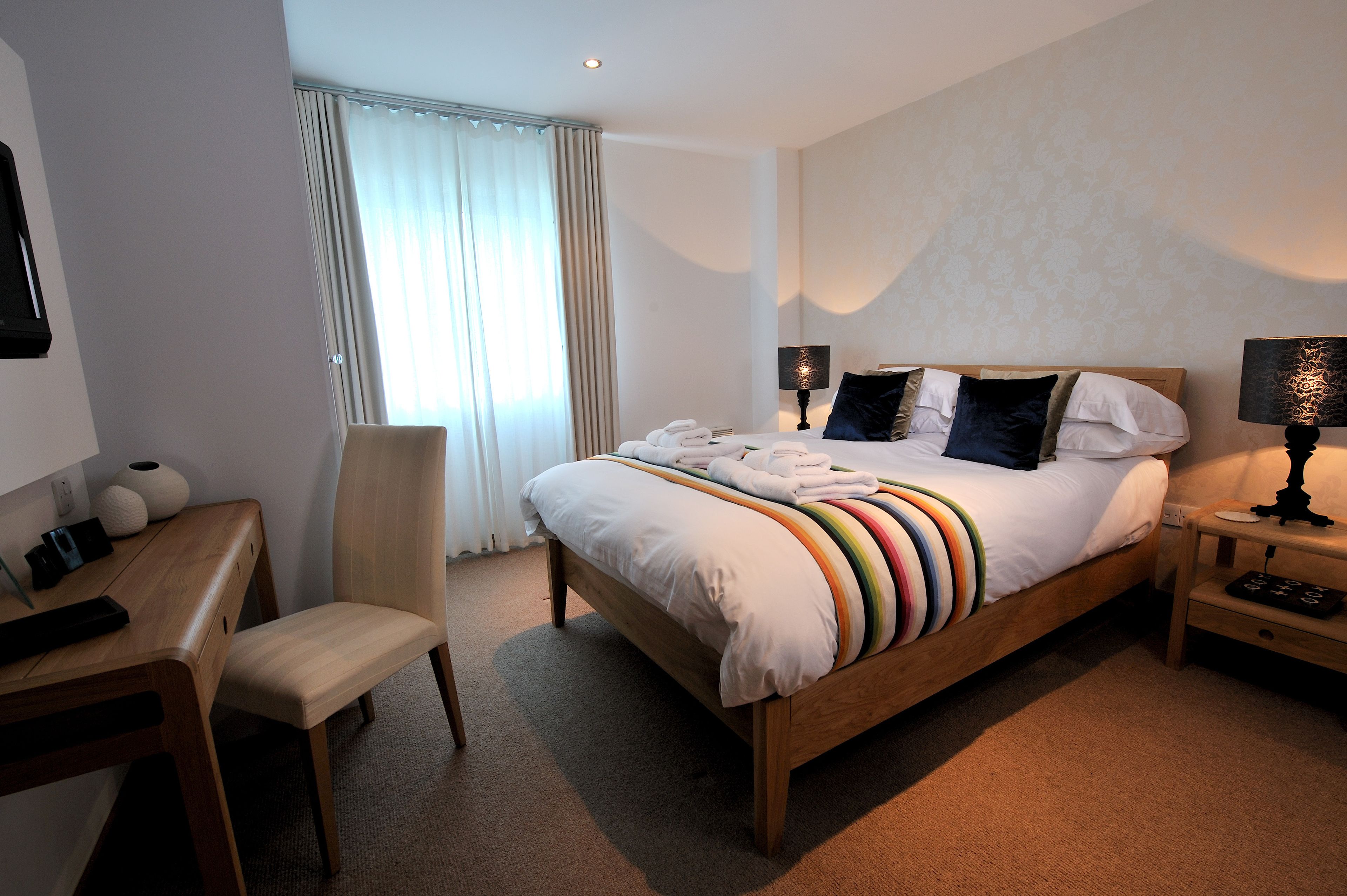 A hotel room with a bed, a desk, a chair, a lamp, and a window with curtains.