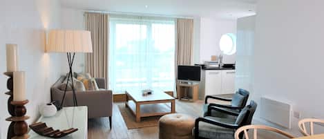 Una sala moderna con sofá, mesa de centro, otomanas y sillas. Hay un televisor, una pequeña área de cocina y un ventanal con cortinas.