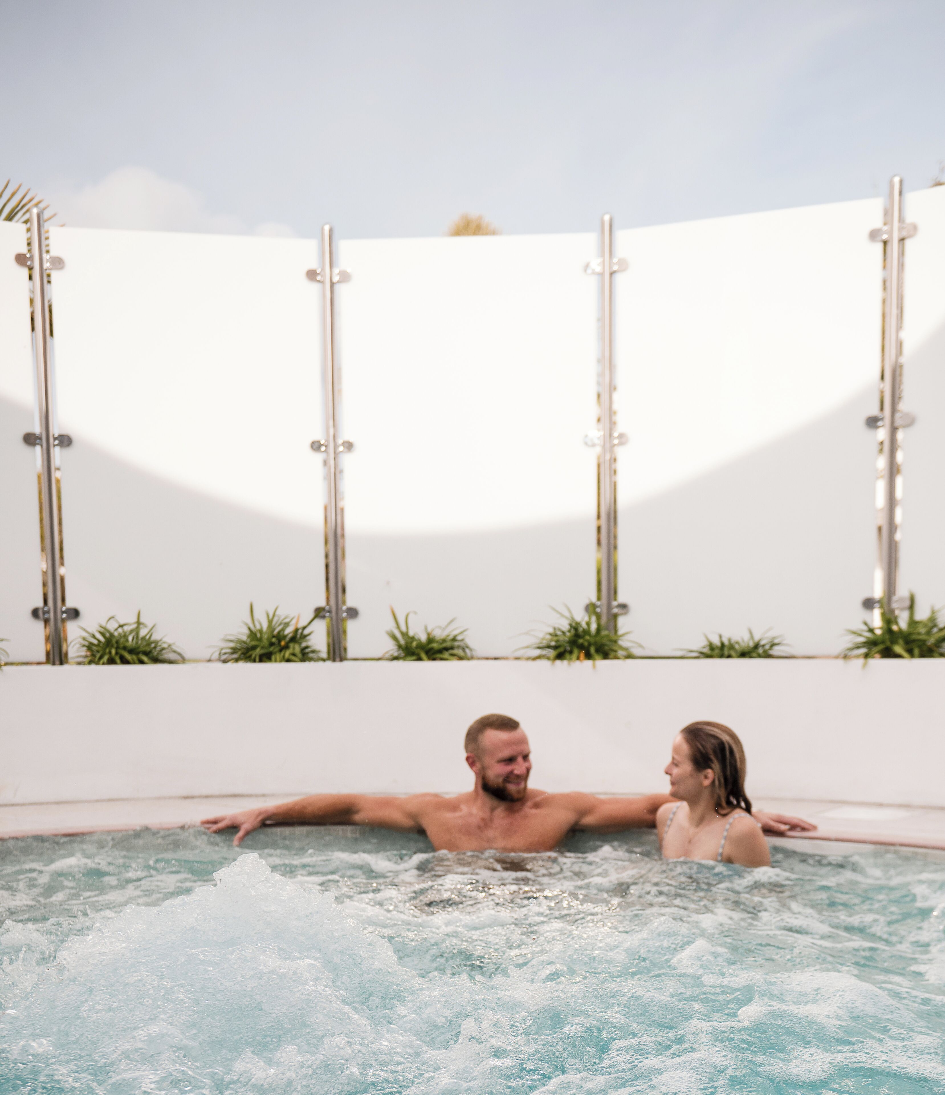 A couple in a hot tub with a white wall and plants in the background.