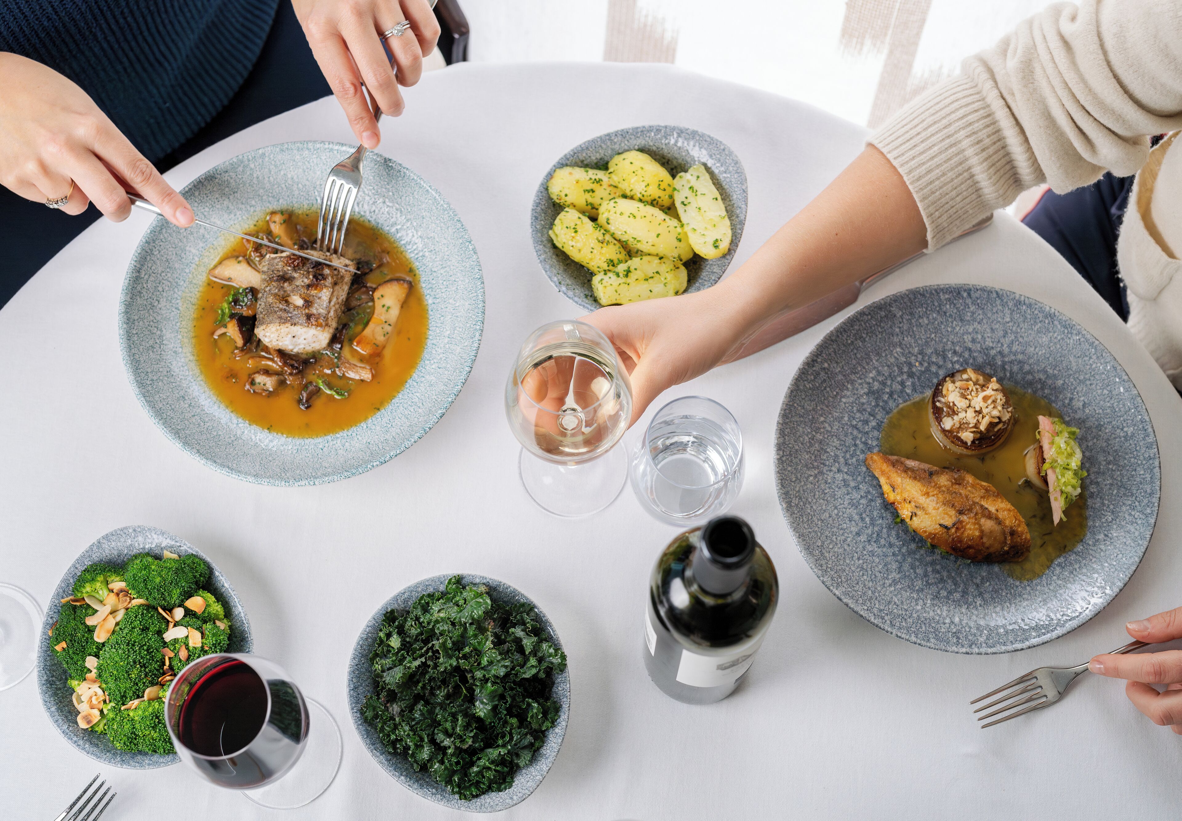 A dining table with various dishes, including a fish dish, potatoes, and greens, along with wine glasses and a bottle.