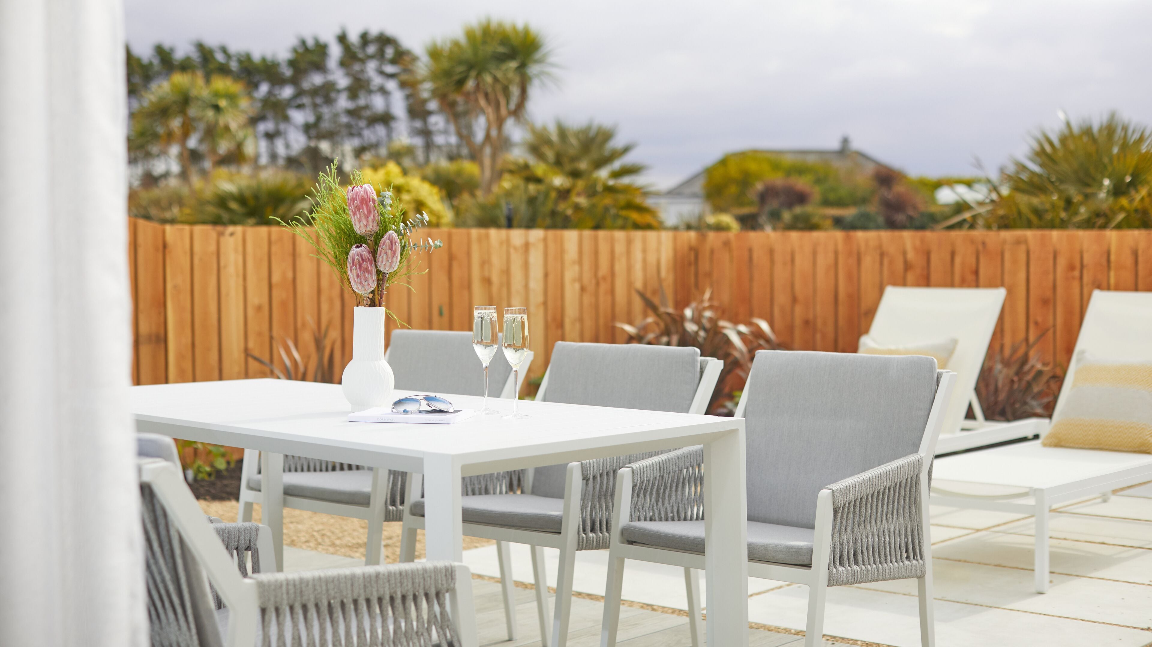 Une terrasse extérieure avec une table blanche, des chaises grises dotées de coussins et une clôture en bois en arrière-plan.