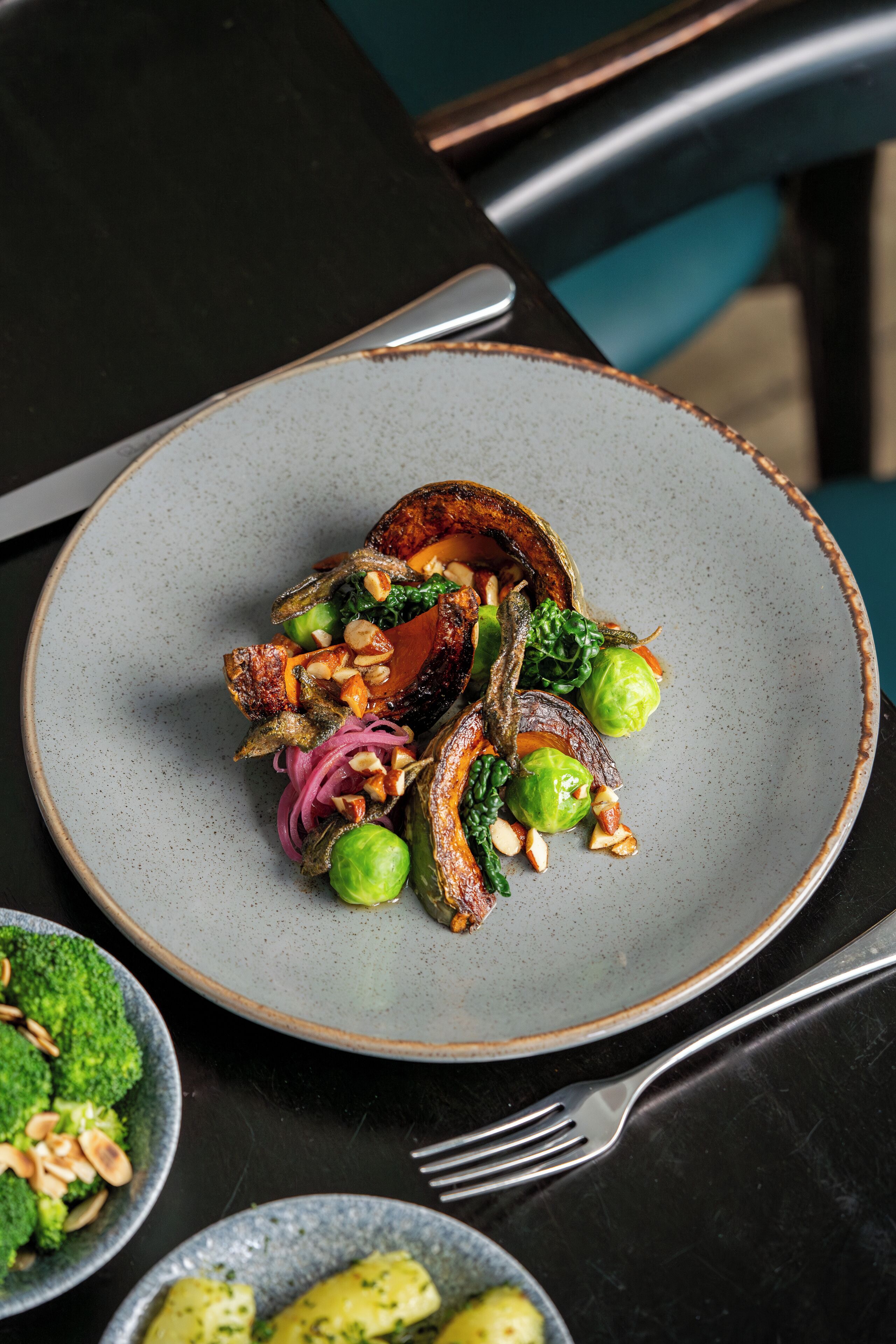 A plate of food with various vegetables and meat, accompanied by a fork and knife on a dark table.