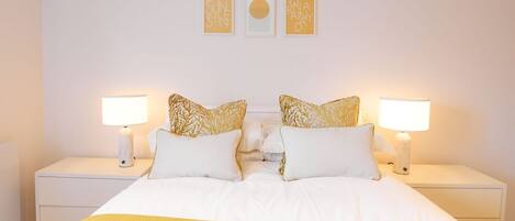 A neatly made bed with white bedding and two decorative pillows, flanked by two bedside tables with lamps, in a simple, clean room.