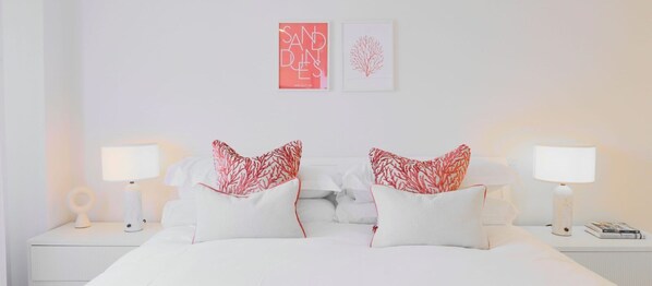 Una cama impecablemente arreglada con ropa de cama blanca y almohadas rojas estampadas, dos lámparas de noche y un cuadro en la pared.