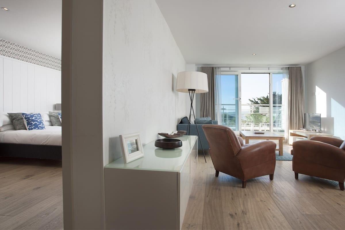 Habitación de hotel moderna con cama, escritorio, dos sillones de cuero, lámpara de pie y vistas al exterior.