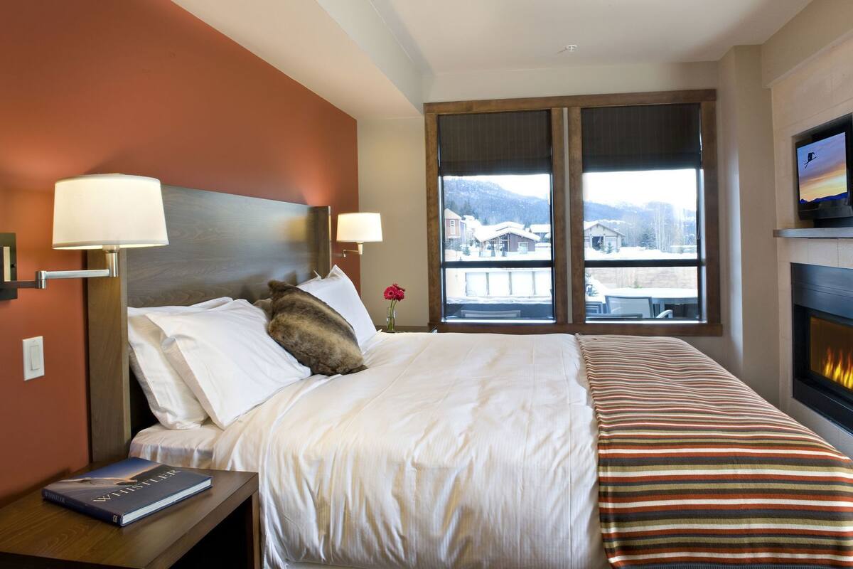 Habitación de hotel con cama, dos lámparas de noche, chimenea, televisor y ventana con vistas a un paisaje nevado.