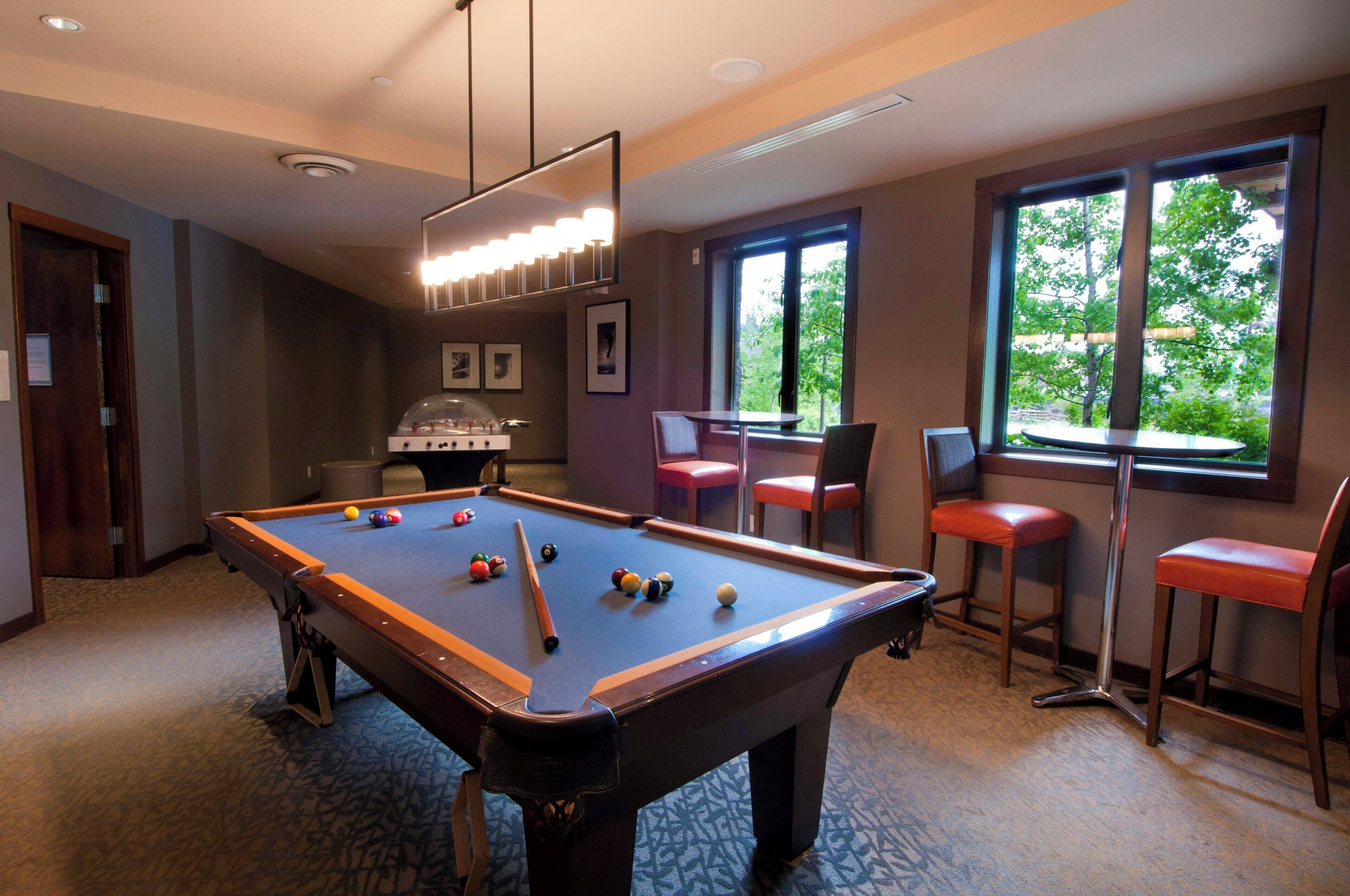 A pool table with balls, a bar area with stools, and a window with a view of trees.