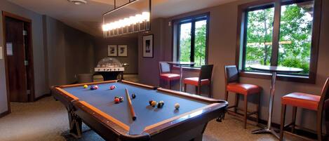 A pool table with balls, a bar area with stools, and a window with a view of trees.