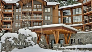 Un bâtiment à plusieurs étages, de style chalet, avec une façade en pierre, des balcons en bois et un espace d’entrée abrité.
