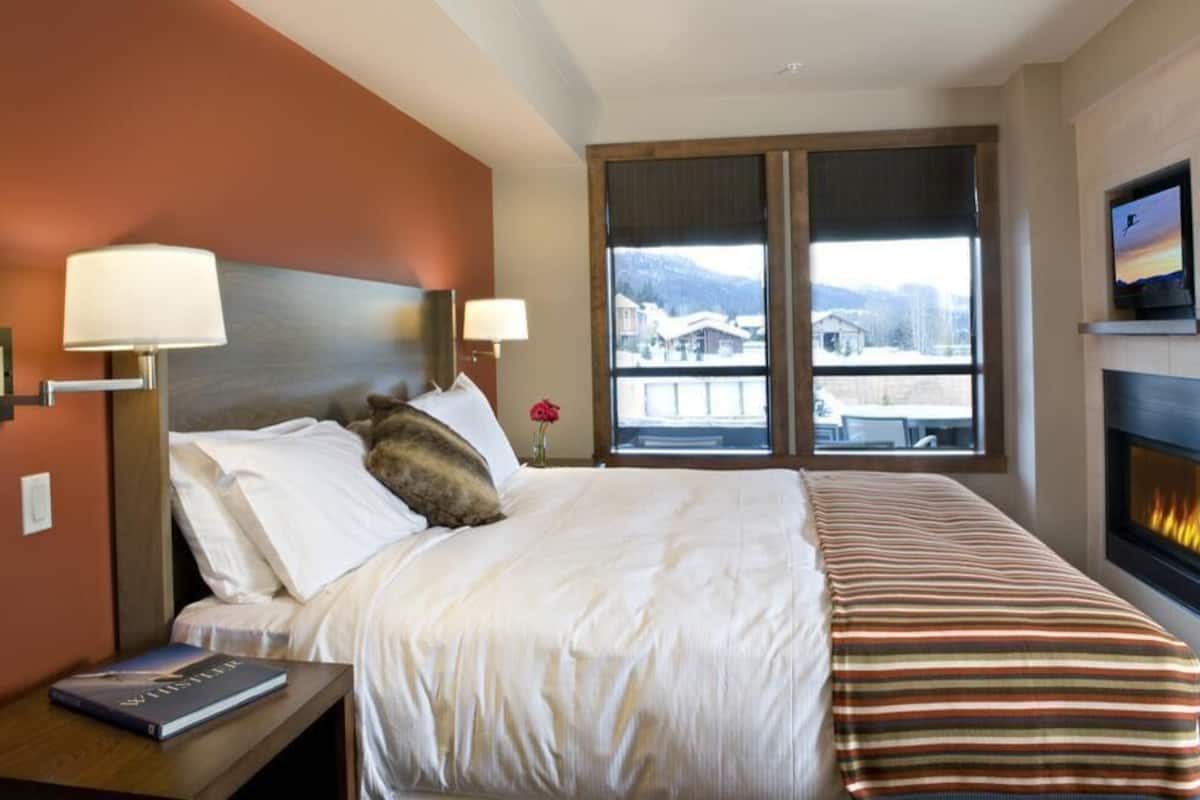 Habitación de hotel con cama, mesita de noche con un libro, televisor, chimenea y una ventana grande con vistas a la montaña.