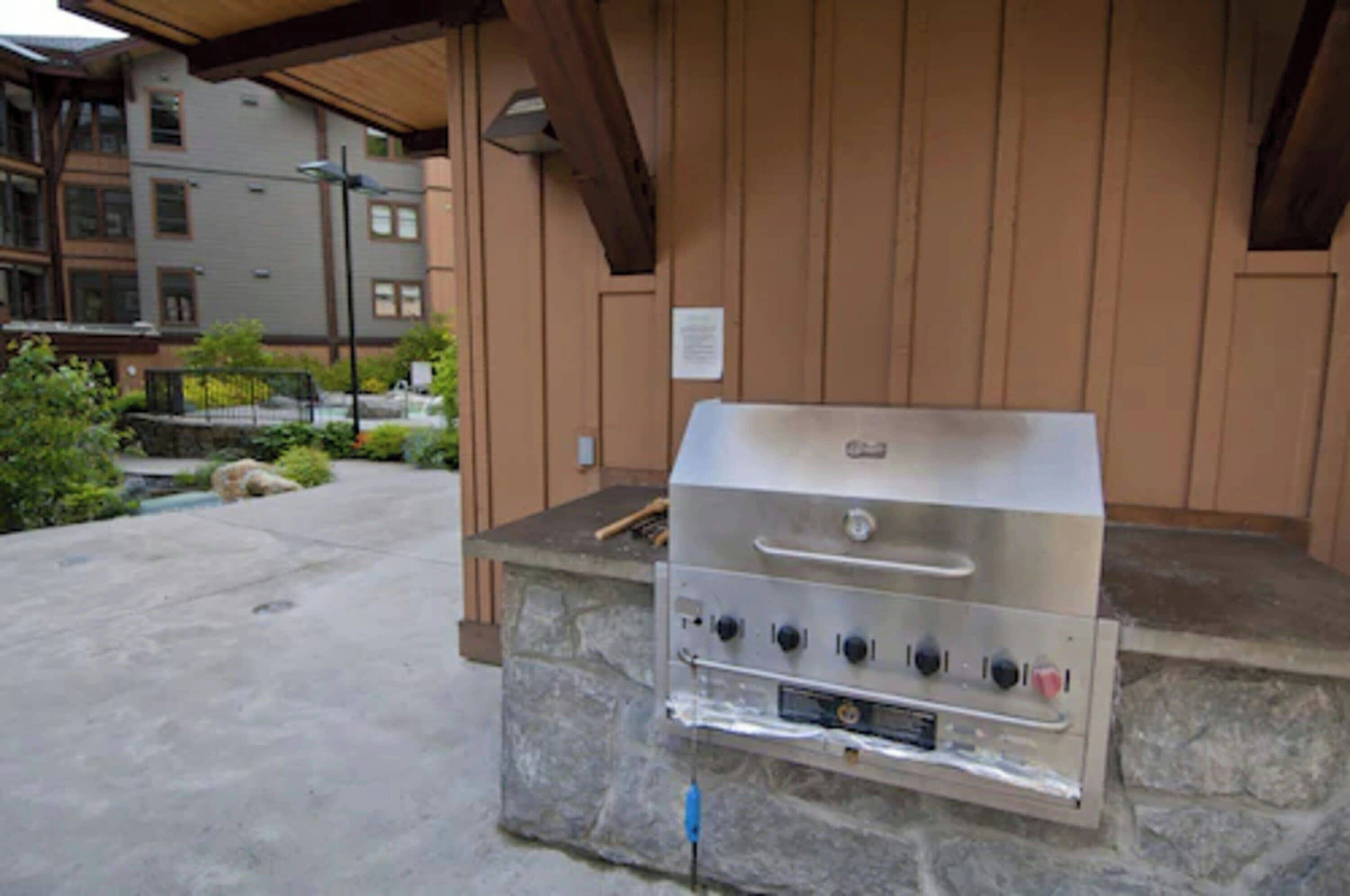 Una parrilla de acero inoxidable con cuatro quemadores, ubicada sobre una plataforma de piedra en un área exterior con una pérgola de madera.